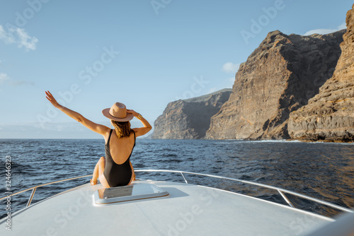 Woman enjoying ocean voyage sitting back on the yacht nose while sailing near the breathtaking rocky coast on a sunset