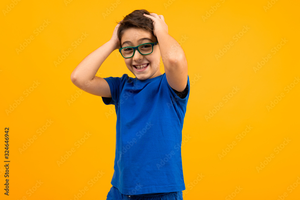 young boy in a blue T-shirt with glasses holds his head on a yellow background with copy space young boy in a blue T-shirt with glasses holds his head on a yellow background with copy space
