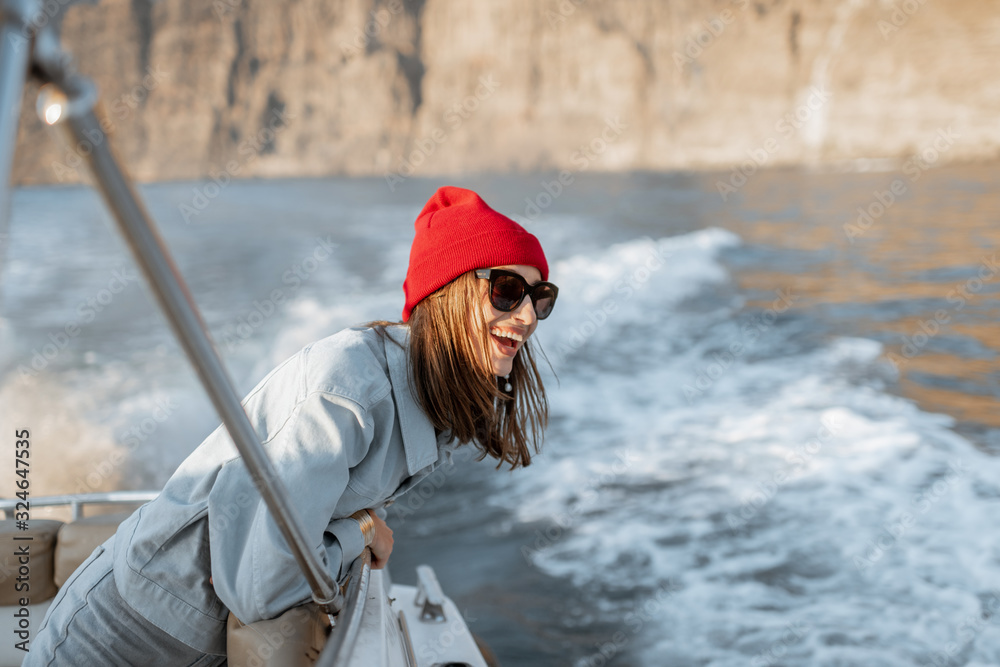Portrait of a young woman traveler dressed casually in red hat and jeans sailing on a yacht near the rocky coast. Concept of a carefree lifestyle and travel Portrait of a young woman traveler dressed casually in red hat and jeans sailing on a yacht near the rocky coast. Concept of a carefree lifestyle and travel
