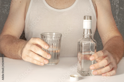 A single man, an alcoholic without a face in a white T-shirt, sits at a table and is about to drink vodka from a faceted glass.