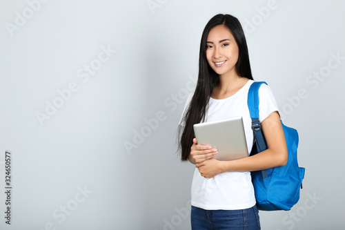Beautiful young woman holding tablet pc and backpack on grey background