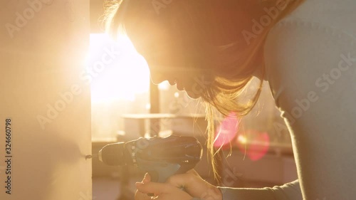 SLOW MOTION, CLOSE UP, LENS FLARE, DOF: Female contractor doing a man's job by using a power drill fasten a wall panel at sunrise. Beautiful Caucasian woman screws small bolts into a gypsum wall board