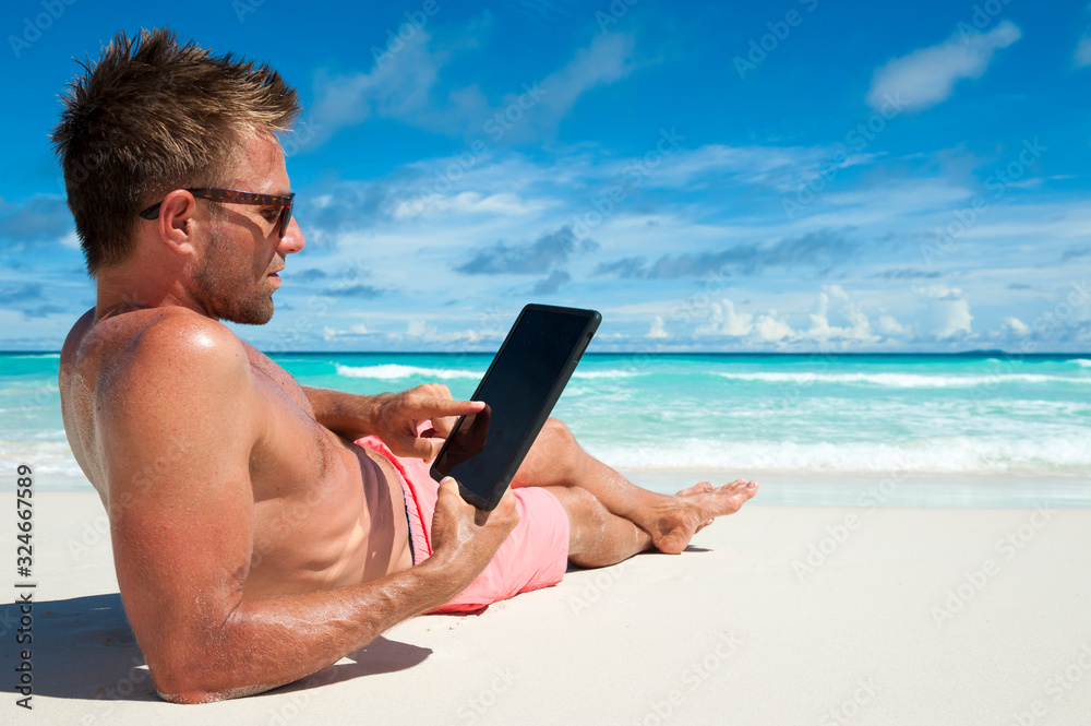 Man relaxing on tropical beach touching the blank screen of his digital tablet computer as waves break on the horizon