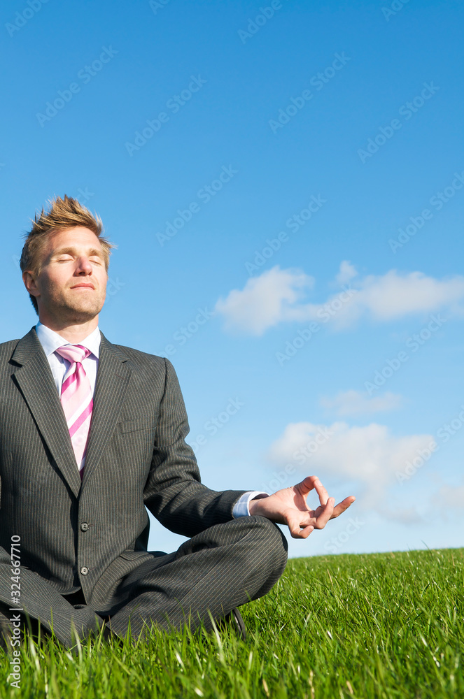 Meditating businessman sitting with eyes closed in a cross-legged yoga pose outdoors in summery green field Meditating businessman sitting with eyes closed in a cross-legged yoga pose outdoors in summery green field
