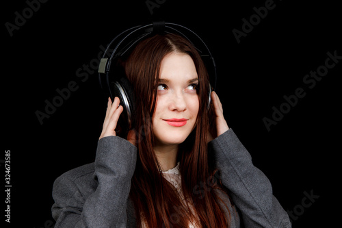 Young pretty woman listening to music in headphones. Girl in grey jacket on black background. Beautiful girl with big earphones © Kateryna