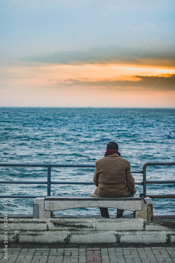 Lonely man sitting alone on the beach at sunset looking away, a ship is pulling away from the horizon Lonely man sitting alone on the beach at sunset looking away, a ship is pulling away from the horizon