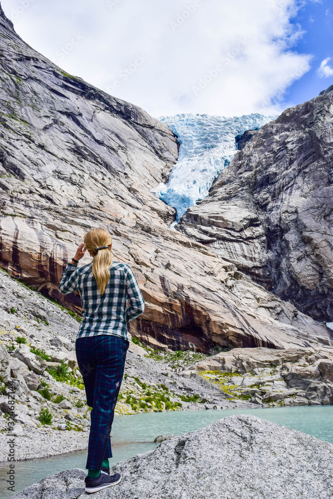 Naklejka premium The young woman near the Briksdalsbreen (Briksdal) glacier, which is the sleeve of large Jostedalsbreen glacier. Melting glacier forms the Briksdalsbrevatnet lake with clear water. Norway.