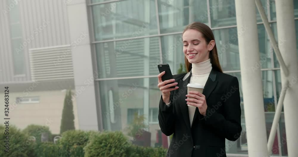 Attractive Smiling female Office Manager stabbing in front of Camera, using Mobile Phone. Big Glass Building in the Background. Drinking Hot Tasty Coffee in her Hands. Typing messages at Screen. Attractive Smiling female Office Manager stabbing in front of Camera, using Mobile Phone. Big Glass Building in the Background. Drinking Hot Tasty Coffee in her Hands. Typing messages at Screen.