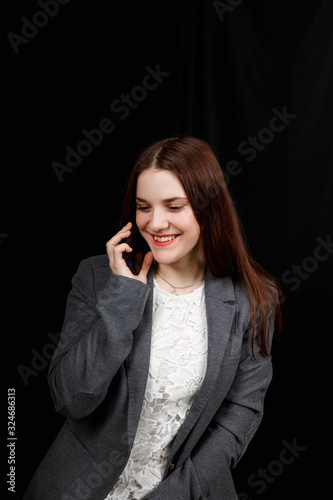 Joyful and successful young fashion blogger is emotionally talking on phone. Beautiful young woman in casual grey jacket is holding smartphone