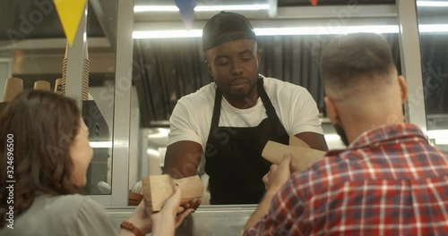 Rear of Caucasian couple standing outdoors at festive food truck cafe window and taking order from African American young vendor. Barman of little street bar giving hot dogs to clients.