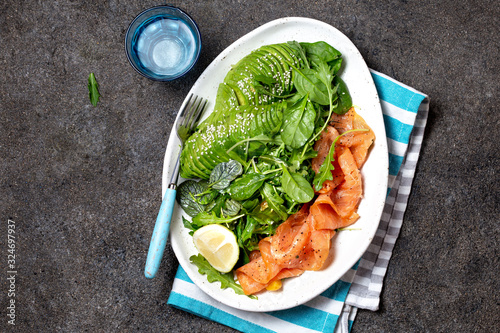 Low carbs salad. Spinach, rucola salad with avocado and salmon. Black concrete background, white plate, top view photo