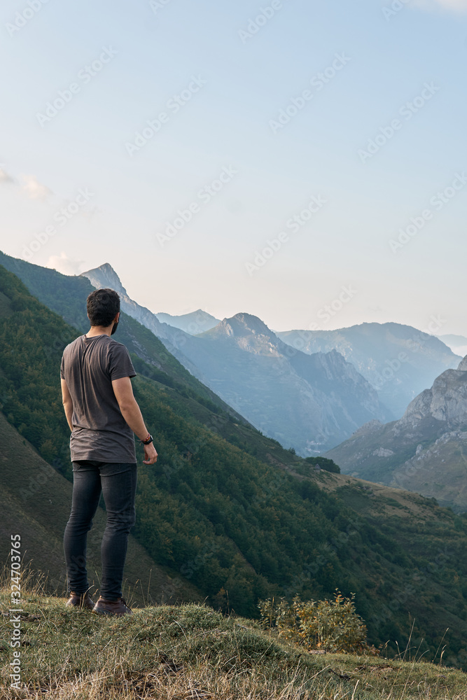 A man contemplates a path that runs through a valley between mountains on a cloudy day with a backpack behind him A man contemplates a path that runs through a valley between mountains on a cloudy day with a backpack behind him
