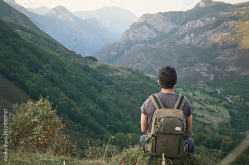A man contemplates a path that runs through a valley between mountains on a cloudy day with a backpack behind him