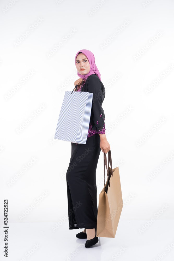 A beautiful Asian female model in a traditional dress kebaya carrying shopping bags isolated on white background. Eidul fitri festive preparation shopping concept. Full length portrait.