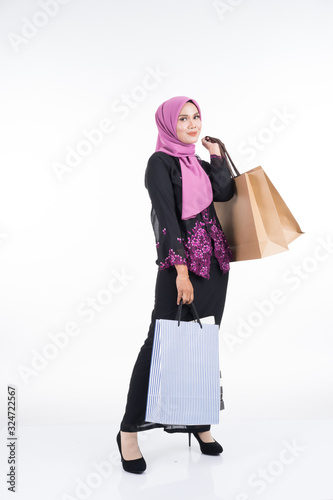 A beautiful Asian female model in a traditional dress kebaya carrying shopping bags isolated on white background. Eidul fitri festive preparation shopping concept. Full length portrait.