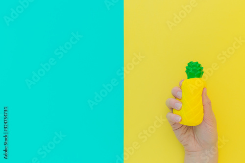 Flat lay antistress toy squish yellow pineapple with green leaves in hand.Bright blue background.Compressing, soft, squeezable items to relieve stress, problems, anxieties, worries. Summer concept