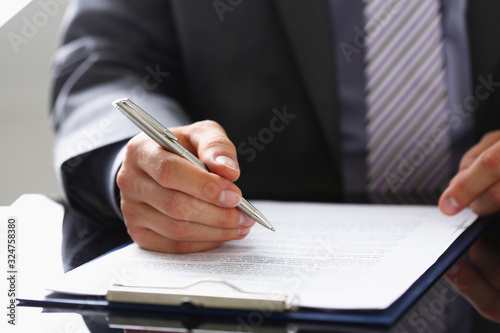 Male arm in suit and tie fill form clipped to pad with silver pen closeup. Sign gesture read pact sale agent bank job make note loan credit mortgage investment finance chief legal law concept