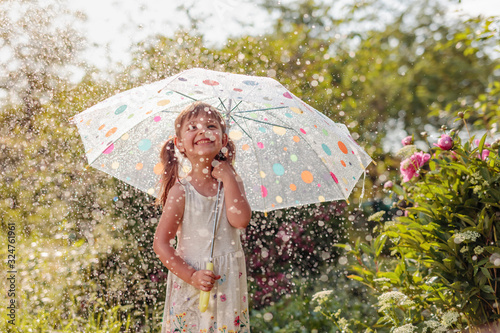 Fotografia Happy little girl in garden under the summer rain with an umbrella