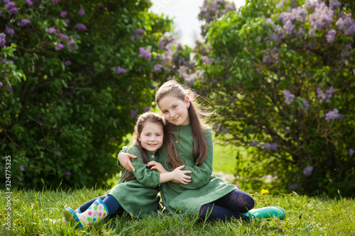 Two sisters in green linen dress have fun in the park with blooming lilacs, enjoy spring and warmth