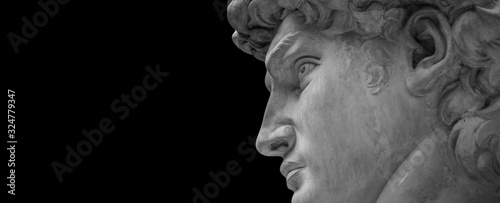 White plaster bust portrait sculpture against a dark background with copyspace, head view. Head and shoulders detail of stone ancient classical statue. Close-up face crop isolated on black