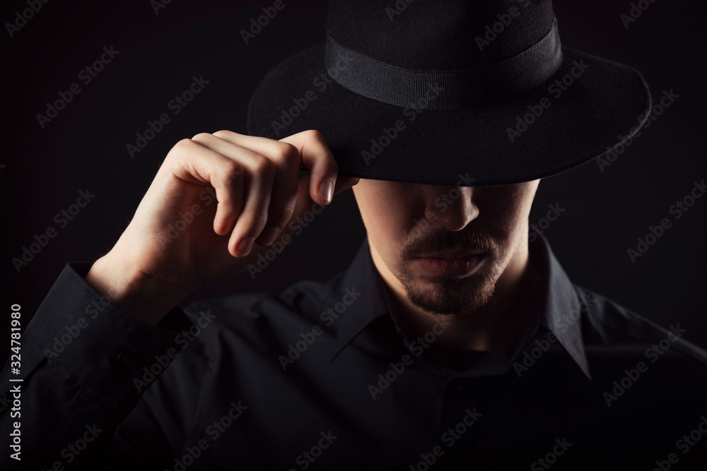 Male with goatee wearing black shirt touching his fedora hat tipped over eyes, dark background