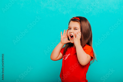Close up portrait of little girl in red clothes is holding hand near her open mouth, screams. isolated on turquoise background with copy space for text 