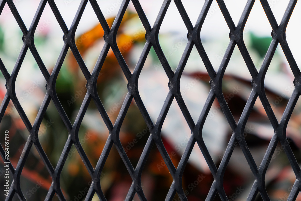 Fototapeta premium Texture of steel diamond shape grill cage fence with unfocused green red and yellow leaves behind it.