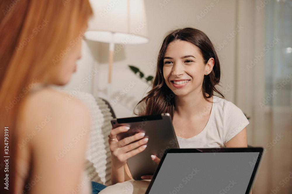 Brunette caucasian girl with a tablet is looking at her red haired friend who is using a laptop sitting on a sofa Brunette caucasian girl with a tablet is looking at her red haired friend who is using a laptop sitting on a sofa