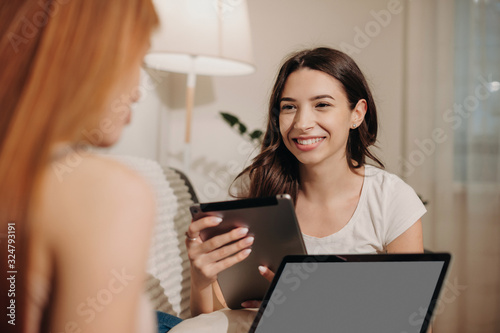 Brunette caucasian girl with a tablet is looking at her red haired friend who is using a laptop sitting on a sofa