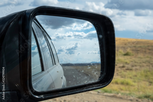 Reflections of cloudy sky, mountains and road in dirty side mirror of suv vehicle.