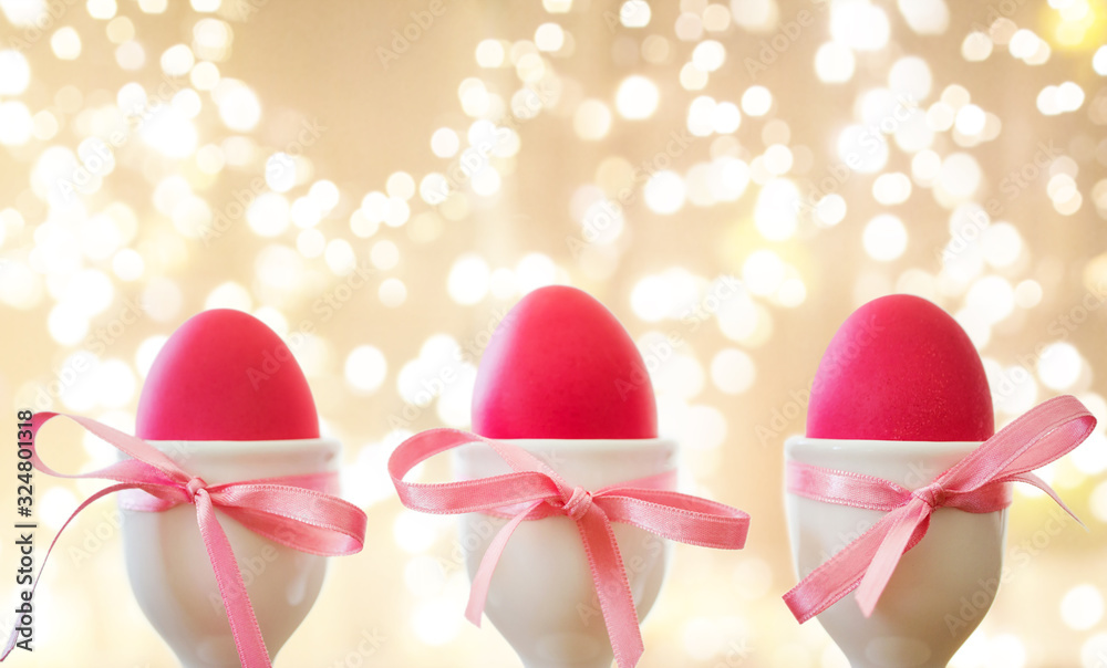 Fototapeta premium easter and holidays concept - pink colored eggs in ceramic cup holders with ribbon over festive lights on beige background