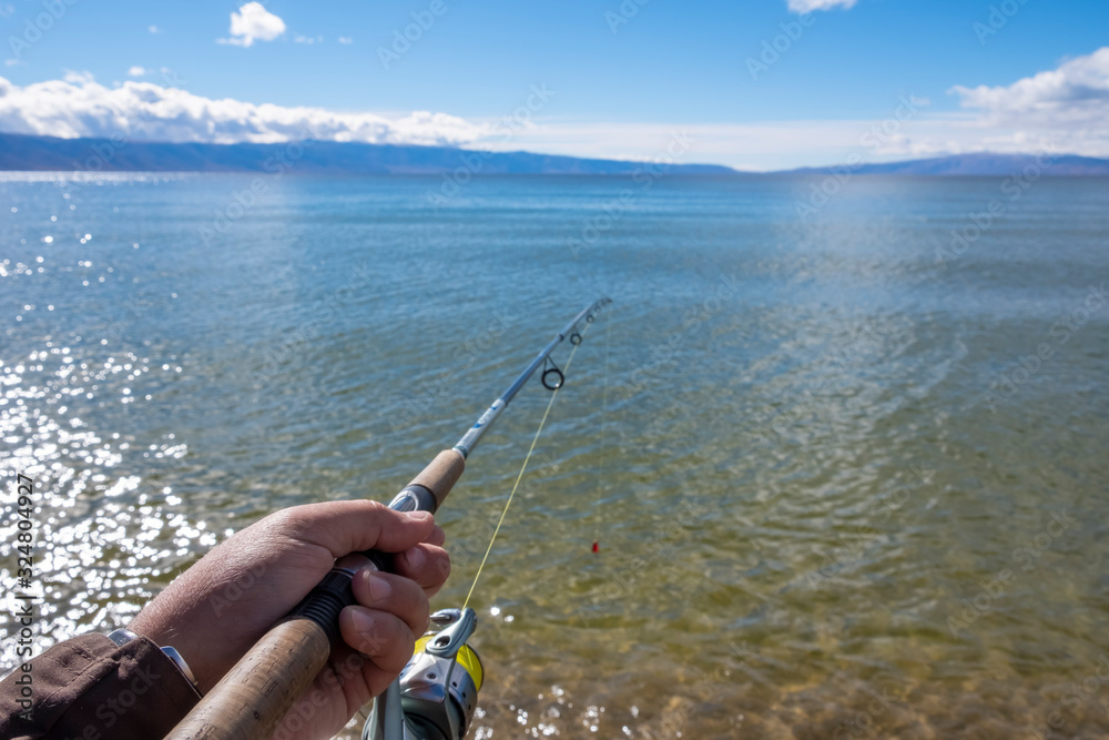 Man's hand is holding a rishing rod with unfocused lake, mountains and sky background. Man's hand is holding a rishing rod with unfocused lake, mountains and sky background.