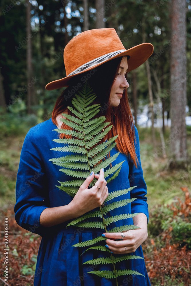 Portrait of beautiful young woman in blue dress and hat with leaf of fern in hands on forest background. Place for text or advertising