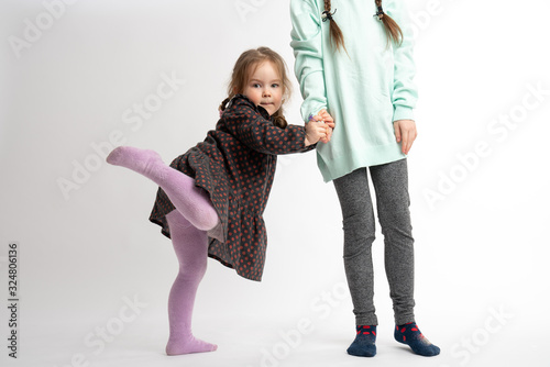 Funny little child girl in dress holds sister's hand and raises her leg, looking at the camera, isolated over white background