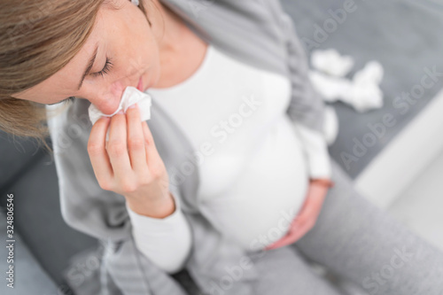 Pregnant woman catching cold, flu, virus, sitting at home on a couch, blowing nose and being sick.