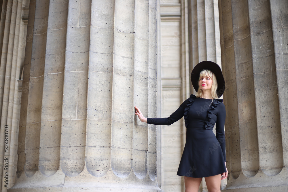 Beautiful long legged blonde in a short black dress and hat is standing near the columns