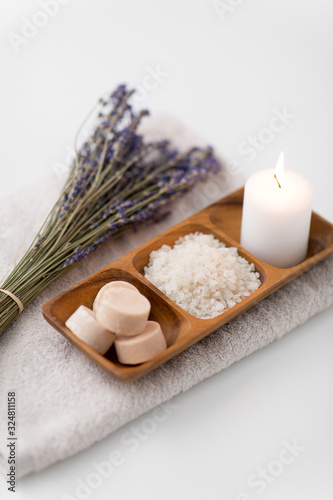 beauty and wellness concept - sea salt  soap and candle on wooden tray and bunch of lavender on bath towel