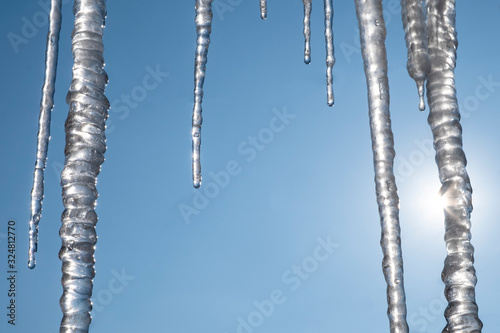 Icicles hanging down from a roof with clear blue sky and sun on a background.