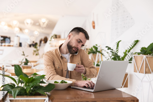 Young handsome bearded smiling man looks busy typing in laptop talking on phone during lunch break at cafe, multitasking concept photo