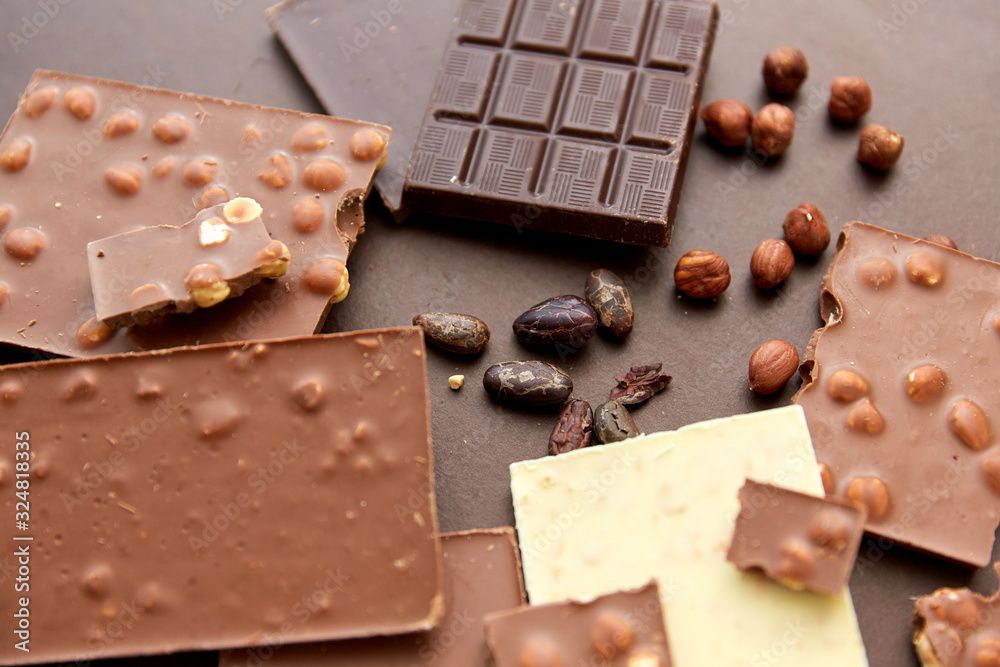 sweets, confectionery and food concept - milk, dark and white chocolate bars with hazelnuts and cocoa beans on brown background sweets, confectionery and food concept - milk, dark and white chocolate bars with hazelnuts and cocoa beans on brown background