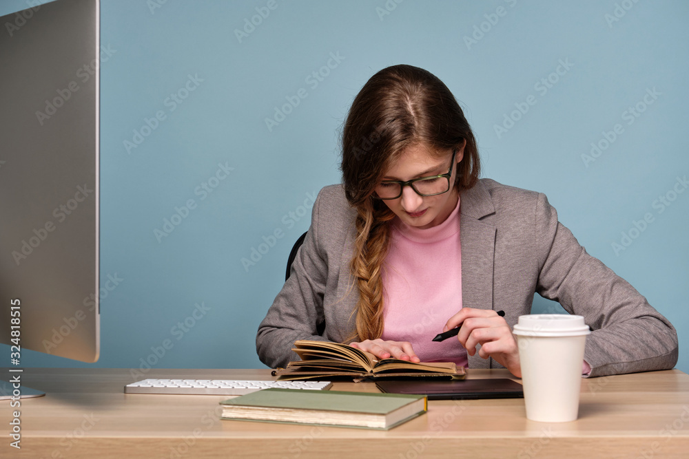 A girl in a gray jacket sits at a table with a computer and carefully looks into a notebook.