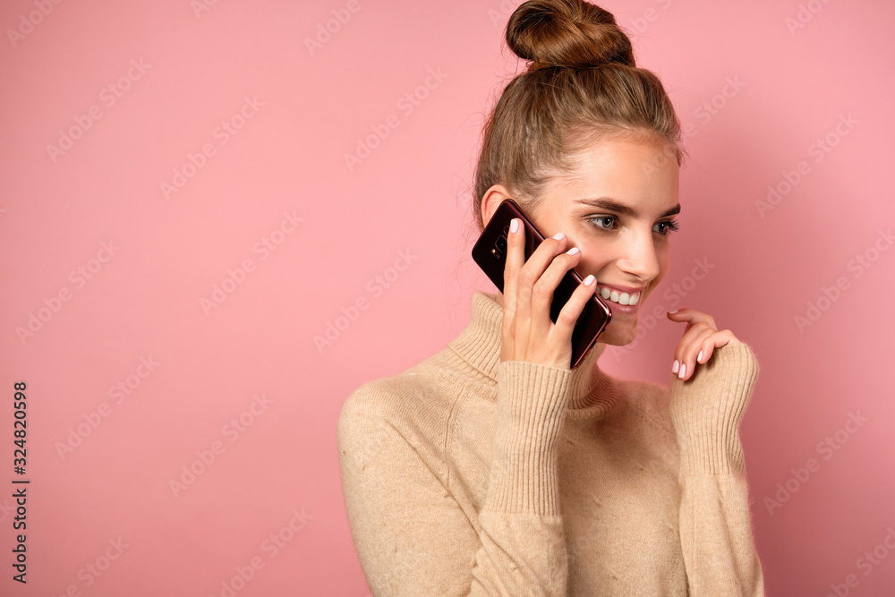 A girl with clean skin and a high bun stands in a half-turn on pink background smiling on the phone. A girl with clean skin and a high bun stands in a half-turn on pink background smiling on the phone.