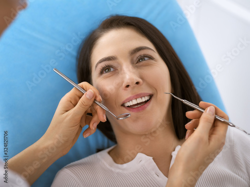 Smiling brunette woman being examined by dentist at dental clinic. Hands of a doctor holding dental instruments near patient's mouth. Healthy teeth and medicine concept