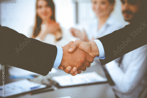 Business people shaking hands at meeting while theirs colleagues clapping and applauding. Group of unknown businessmen and women in modern white office. Success teamwork, partnership and handshake