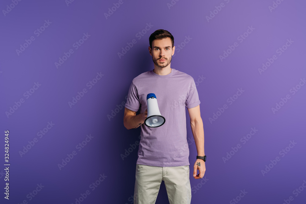 serious young man holding megaphone and looking at camera on purple background