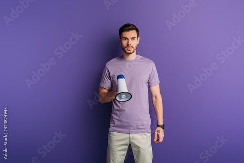 serious young man holding megaphone and looking at camera on purple background © LIGHTFIELD STUDIOS