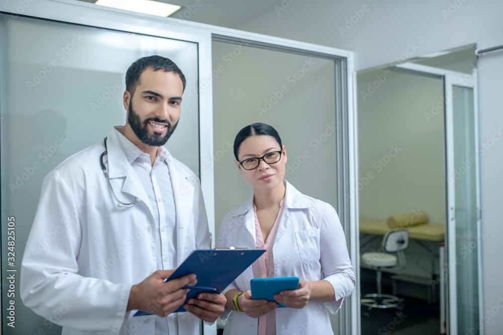 Dark-haired male and female doctors standing in the room together Dark-haired male and female doctors standing in the room together