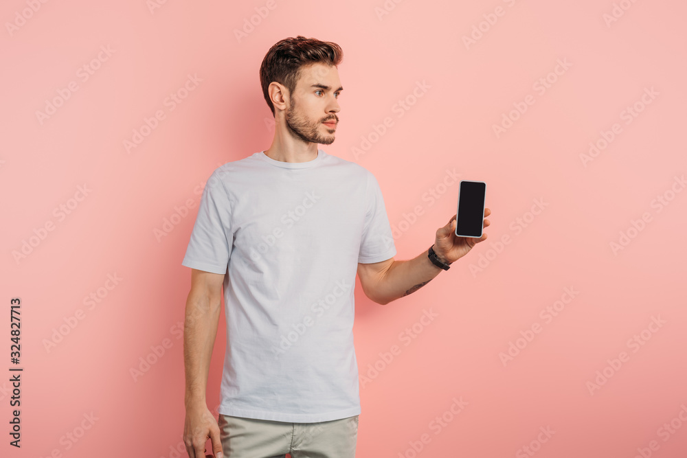 confident young man showing smartphone with blank screen and looking away on pink background confident young man showing smartphone with blank screen and looking away on pink background