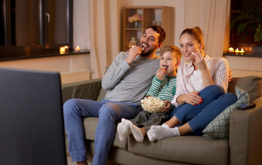 family, leisure and people concept - happy smiling father, mother and little son watching tv and eating popcorn at home at night