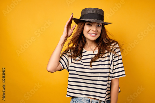A beautiful brunette in a wicker hat and with flying hair stands on a yellow background and smiles charmingly.